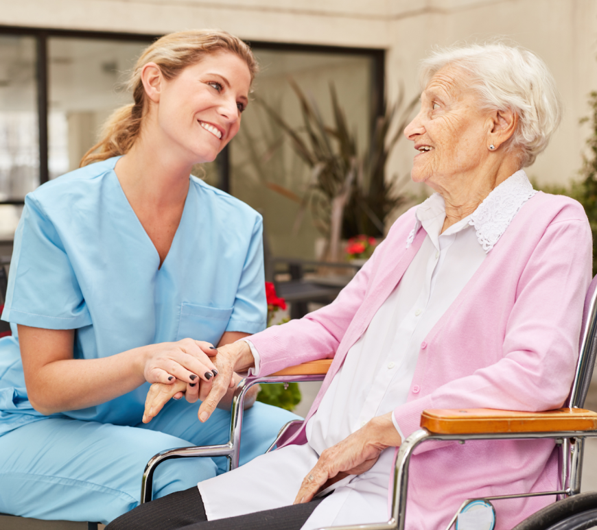 Outside, an elderly white woman sits in a wheelchair. On a bench next to her is a younger white woman wearing scrubs smiling and holding her hand.
